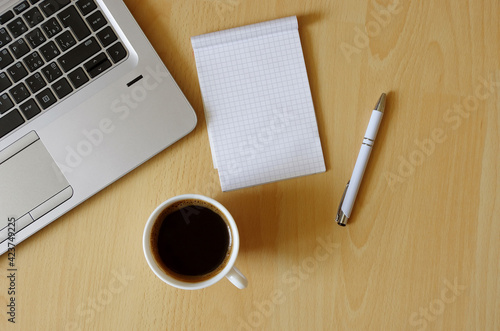 Laptop with coffee cup, notebook and pen on wooden background Top view. Concept for home office. Business concept. Work from home. Desk with laptop, blank notepad, coffee cup and pen on wooden table.