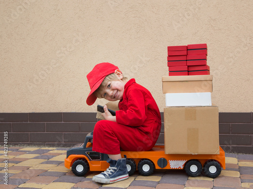 happy toddler boy in a red overalls and a cap sits on a large toy car loaded with parcels - boxes. games for kids - postman, be like dad photo