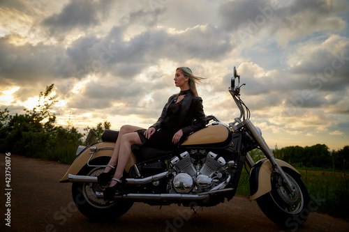 Attractive female brunette motorcyclist with motorcycle in a summer evening during sunset. Beautiful girl on nature and sky with sun and clouds background. Adventure concept