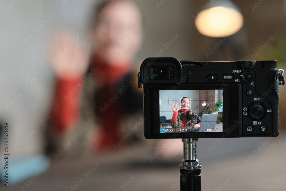 Portrait of young woman waving at camera while sitting at the table with laptop and recording herself on camera Portrait of young woman waving at camera while sitting at the table with laptop and recording herself on camera