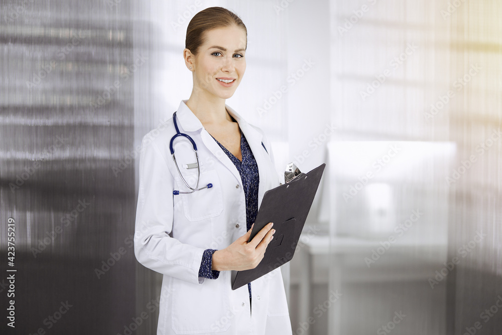 Cheerful female doctor standing in sunny clinic. Portrait of friendly smiling woman-physician. Medicine concept Cheerful female doctor standing in sunny clinic. Portrait of friendly smiling woman-physician. Medicine concept