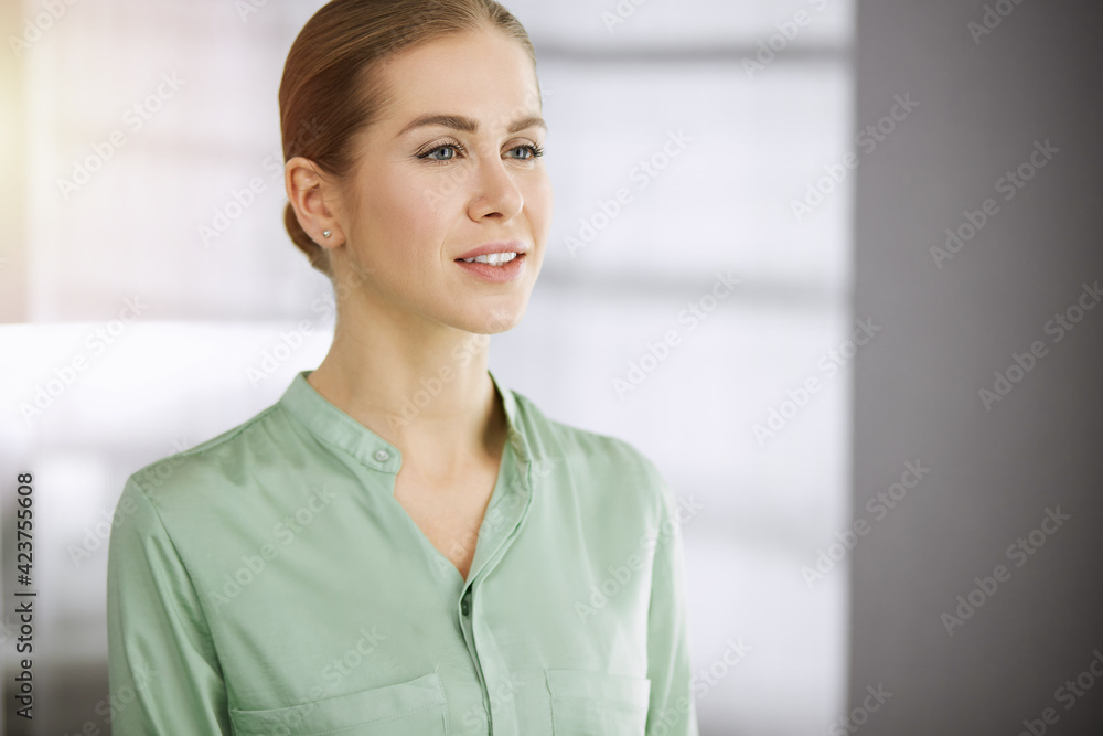 Beautiful adult business woman dressed in green blouse standing straight in sunny office. Business headshot
