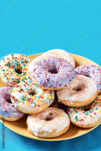 Light and fluffy dessert. Close up shot of different colourful round glazed donuts with sprinkles on yellow plate over blue background