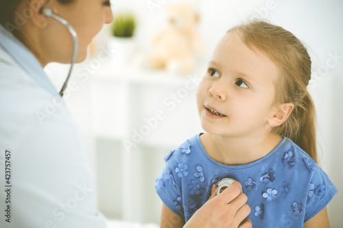 Doctor examining a child by stethoscope. Happy smiling girl patient dressed in blue dress is at usual medical inspection. Medicine concept
