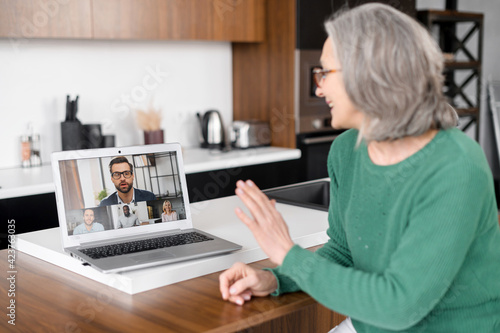 Mature female holding video conference on the laptop sitting at home, senior woman involved virtual meeting with colleagues, watching online video webinar