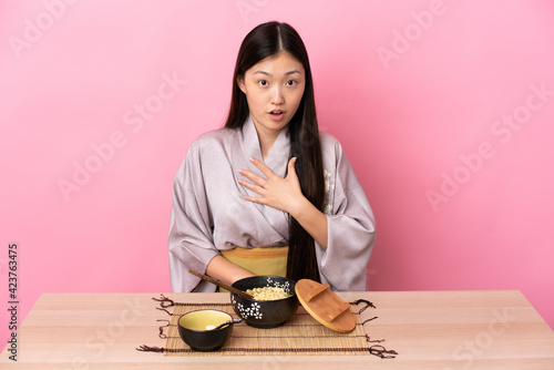Young Chinese girl wearing kimono and eating noodles surprised and shocked while looking right