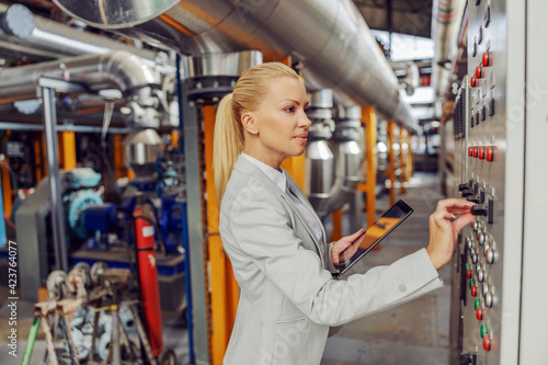 Female blond supervisor standing in heating plant next to dashboard, adjusting settings and holding tablet.