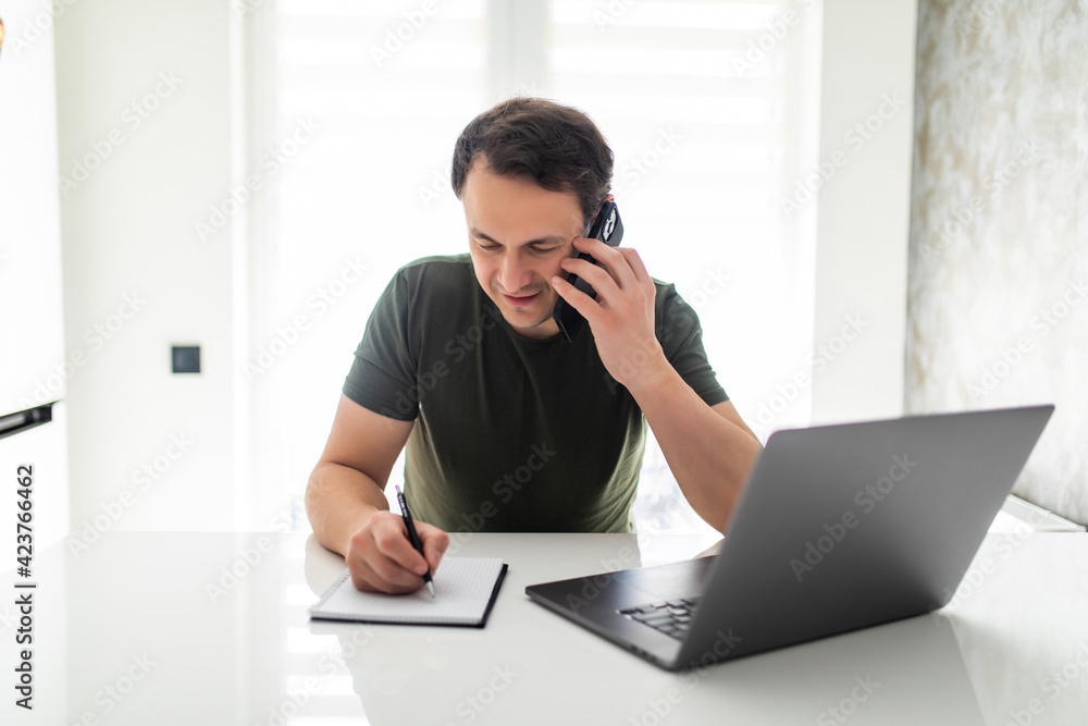 Handsome young man calculate bills make notice using laptop and talking on cellphone in kitchen. Handsome young man calculate bills make notice using laptop and talking on cellphone in kitchen.
