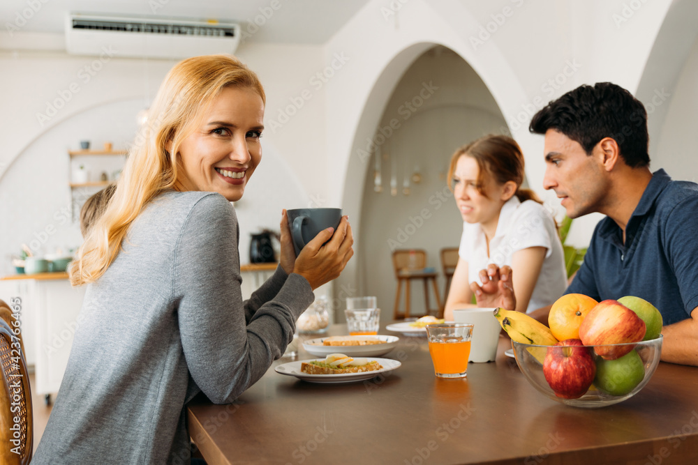 Portrait of beautiful young mother holding cup of coffee while smiling and looking at camera with handsome husband and children in a conversation during breakfast