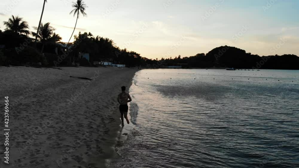 Following a young fast man running on the sand beach in Thailand. Aerial view of a jogger on a tropical beach at sunset Following a young fast man running on the sand beach in Thailand. Aerial view of a jogger on a tropical beach at sunset