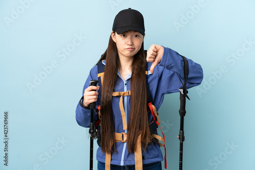 Young Chinese girl with backpack and trekking poles over isolated blue background showing thumb down with negative expression © luismolinero