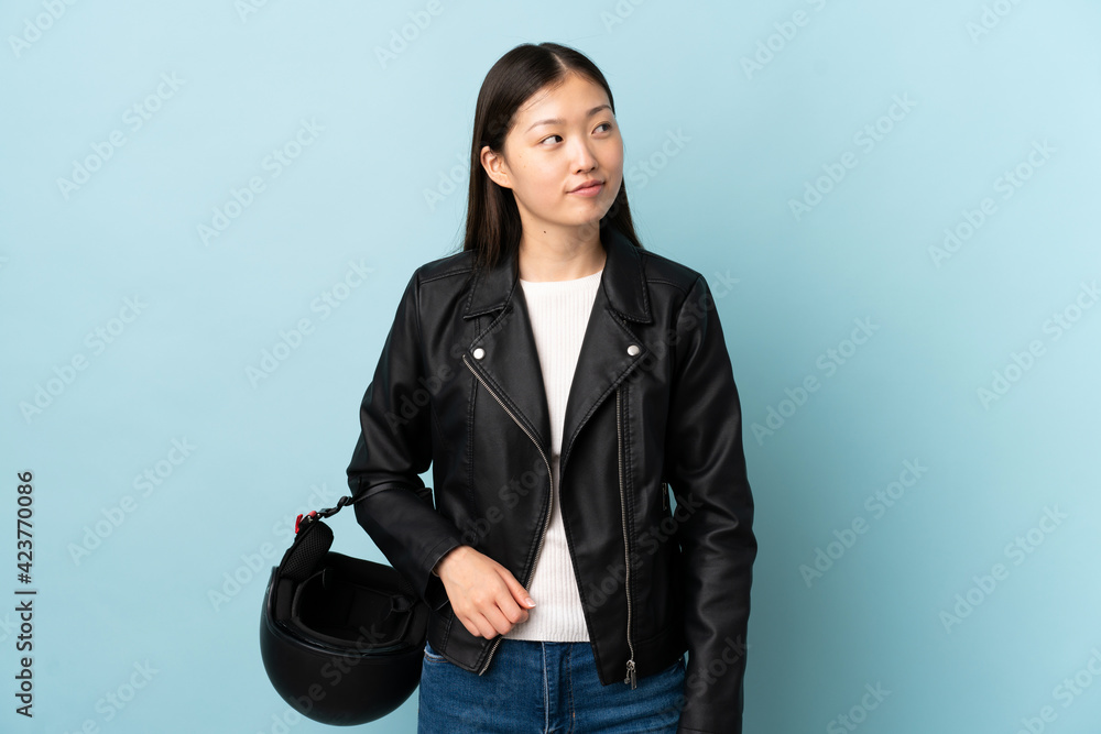 Chinese woman holding a motorcycle helmet over isolated blue background standing and looking to the side Chinese woman holding a motorcycle helmet over isolated blue background standing and looking to the side