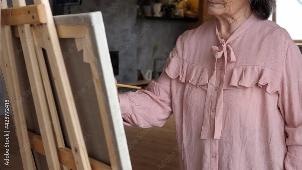 Elderly woman artist in an elegant blouse paints a picture on canvas in an art studio. Home leisure, retired hobbies, hobbies, lifelong work Elderly woman artist in an elegant blouse paints a picture on canvas in an art studio. Home leisure, retired hobbies, hobbies, lifelong work