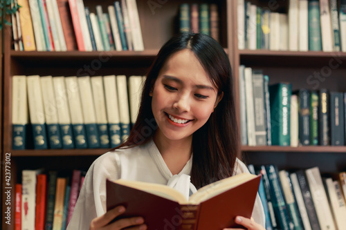 Happy Asian young knowledgable business woman reading the book in library with various book with books on the bookshelf background