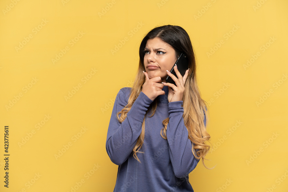 Young Russian girl using mobile phone isolated on yellow background having doubts Young Russian girl using mobile phone isolated on yellow background having doubts
