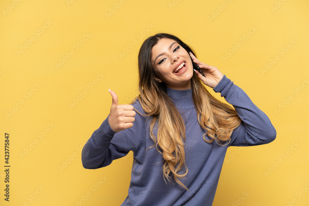 Young Russian girl using mobile phone isolated on yellow background with thumbs up because something good has happened Young Russian girl using mobile phone isolated on yellow background with thumbs up because something good has happened