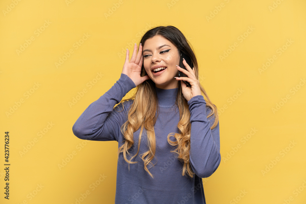 Young Russian girl using mobile phone isolated on yellow background listening to something by putting hand on the ear Young Russian girl using mobile phone isolated on yellow background listening to something by putting hand on the ear