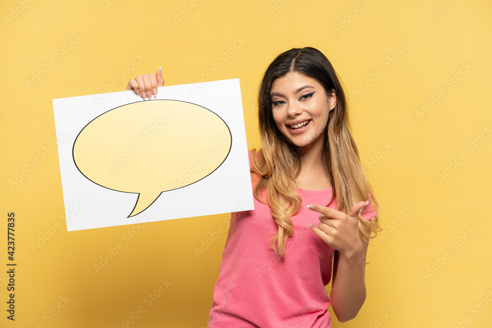 Young Russian girl isolated on yellow background holding a placard with speech bubble icon and pointing it Young Russian girl isolated on yellow background holding a placard with speech bubble icon and pointing it
