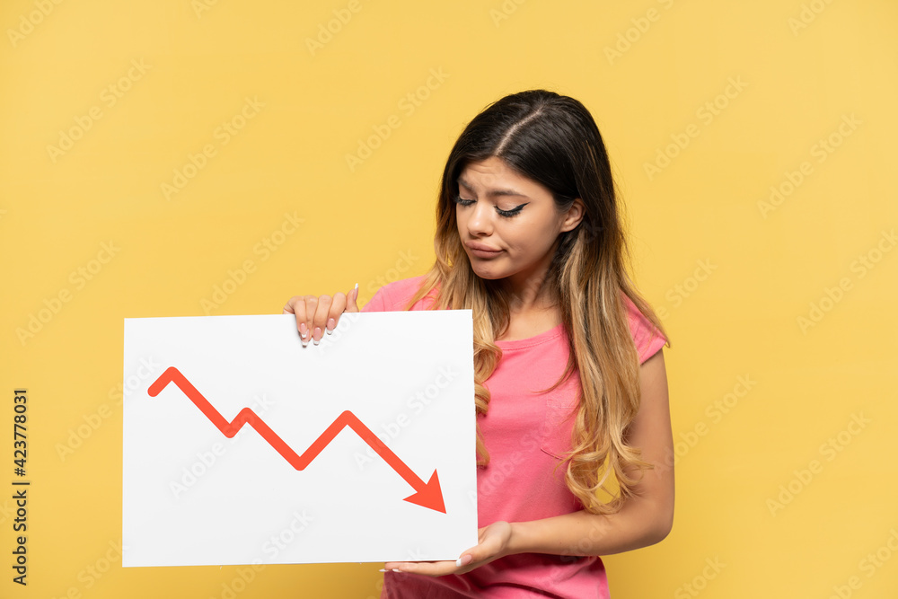 Young Russian girl isolated on yellow background holding a sign with a decreasing statistics arrow symbol with sad expression Young Russian girl isolated on yellow background holding a sign with a decreasing statistics arrow symbol with sad expression