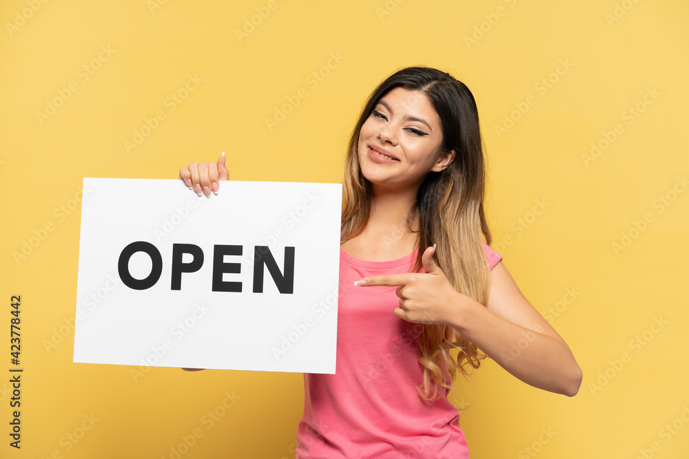 Young Russian girl isolated on yellow background holding a placard with text OPEN and pointing it Young Russian girl isolated on yellow background holding a placard with text OPEN and pointing it