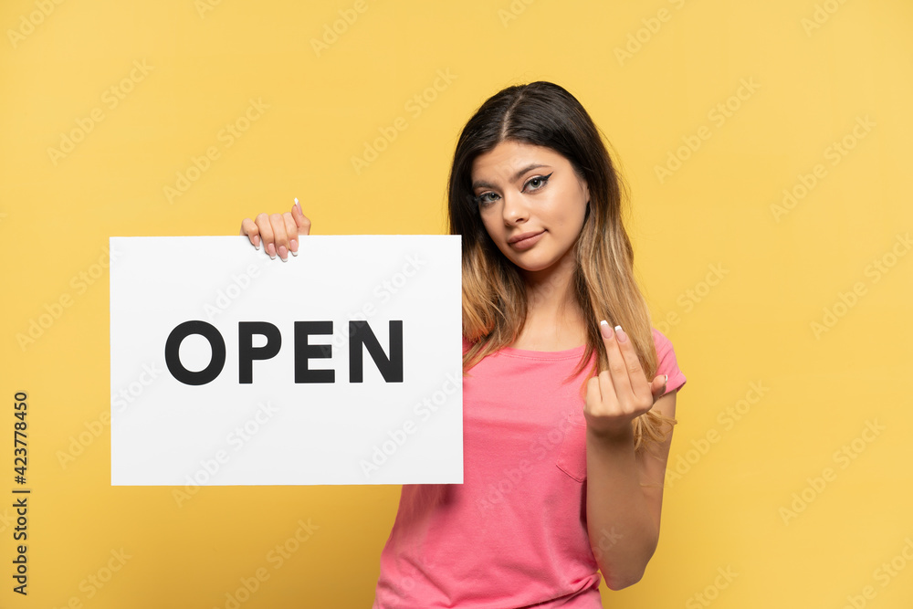 Young Russian girl isolated on yellow background holding a placard with text OPEN and doing coming gesture Young Russian girl isolated on yellow background holding a placard with text OPEN and doing coming gesture