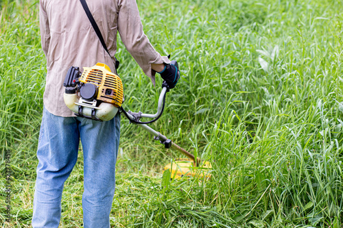 a man in a rumpled shirt and jeans mows the long grass on a suburban plot with a gasoline lawn mower in the summer afternoon