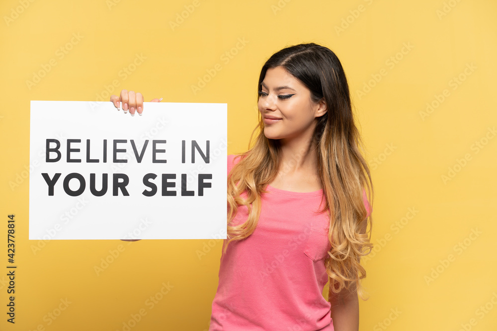 Young Russian girl isolated on yellow background holding a placard with text Believe In Your Self Young Russian girl isolated on yellow background holding a placard with text Believe In Your Self