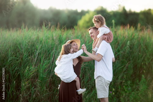 Stylish charming Caucasian young family with two children hugging in summer in a green park, baby girl blond with Down syndrome with parents and younger sister, toning, evening light