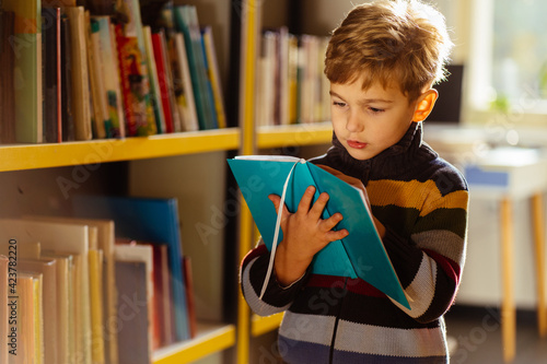 Child s brain development, learn to read, cognitive skills concept. Preschool boy reading book in library next to bookshelves, caucasian blond kid boy is concentrated on education, getting knowledge. photo