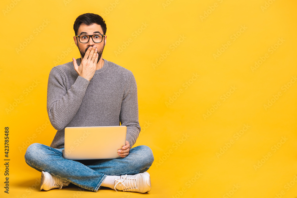 Young bearded man shocked surprised amazed with laptop computer. Funny image of young Caucasian male student model sitting on floor isolated on yellow background. Young bearded man shocked surprised amazed with laptop computer. Funny image of young Caucasian male student model sitting on floor isolated on yellow background.