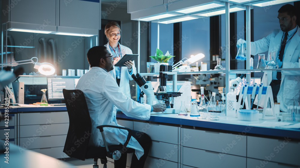 Female and Male Medical Research Scientists Have a Conversation While Conducting Experiments Under a Microscope, Writing Analysis Results on a Tablet Computer. Modern Biological Science Laboratory. Female and Male Medical Research Scientists Have a Conversation While Conducting Experiments Under a Microscope, Writing Analysis Results on a Tablet Computer. Modern Biological Science Laboratory.