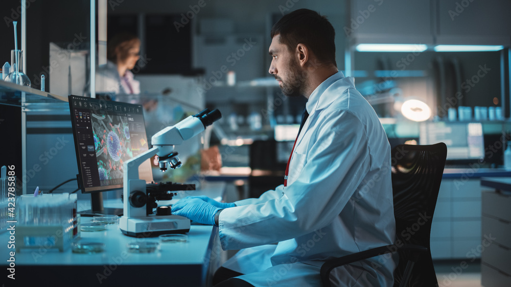 Medical Research Scientist Conducts DNA Experiments Under a Microscope and Writes Results on Desktop Computer in a Biological Science Laboratory. Handsome Caucasian Lab Engineer in White Coat.
