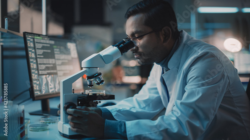 Medical Research Scientist Conducts DNA Experiments Under a Digital Microscope in a Biological Applied Science Laboratory. Multiethnic Lab Engineer in Glasses in White Coat Working on Vaccine.