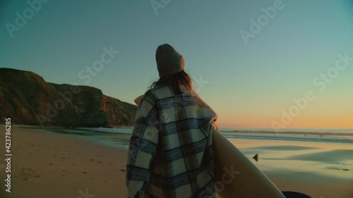 Authentic and real female surfer in casual clothes walks to surfing spot on winter beach. Early sunrise at epic travel surf location. Millennial trendy and cool young woman ready for cold water