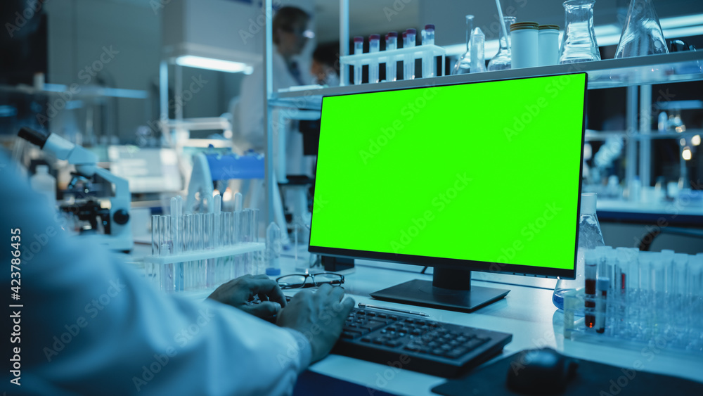 Medical Research Scientist Working on Desktop Computer with Green Screen Mock Up Template in Applied Science Research Laboratory. Lab Engineers in White Coats Conduct Experiments in the Background. Medical Research Scientist Working on Desktop Computer with Green Screen Mock Up Template in Applied Science Research Laboratory. Lab Engineers in White Coats Conduct Experiments in the Background.