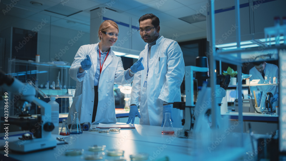 Female and Male Medical Research Scientists Have Conversation and Use Futuristic Virtual Interface with Gestures in a Modern Science Laboratory. Space for Augmented Reality Hologram is Left Empty. Female and Male Medical Research Scientists Have Conversation and Use Futuristic Virtual Interface with Gestures in a Modern Science Laboratory. Space for Augmented Reality Hologram is Left Empty.