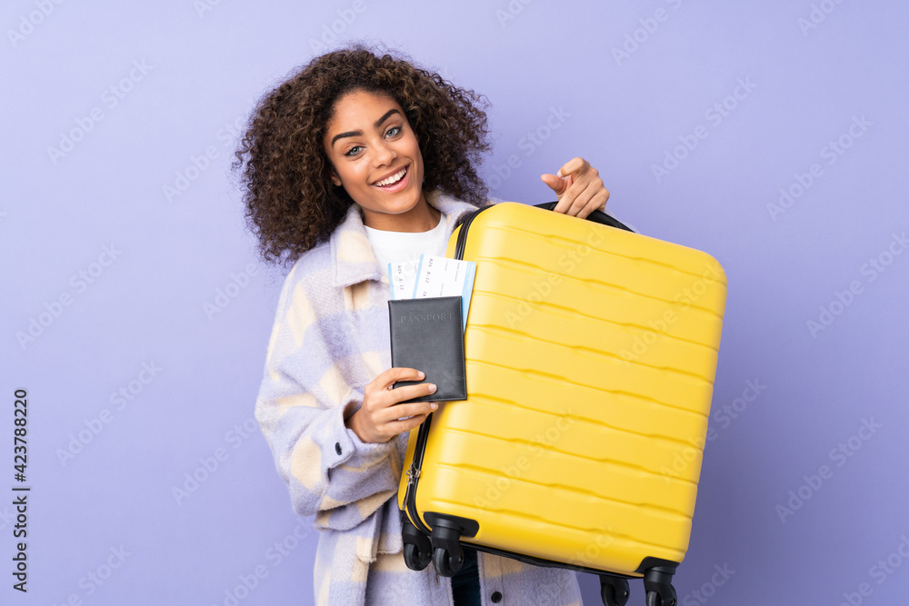 Fototapeta premium Young African American woman isolated on purple background in vacation with suitcase and passport