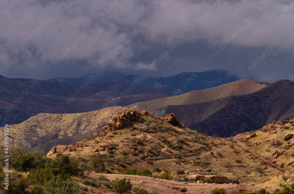 Moody and stormy skies dominate the scene with sun breaking through to light the desert foreground. 