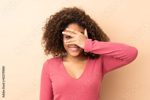Young African American woman isolated on beige background covering eyes by hands and smiling