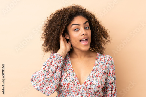 Young African American woman isolated on beige background listening to something by putting hand on the ear