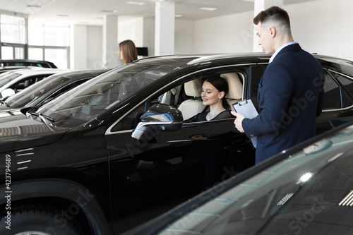 Side view of young beautiful woman sitting inside car and holding hand on steering wheel. She smiling and talking with manager of car dealership. Car agent representing inside of automobile. © Artem Zakharov