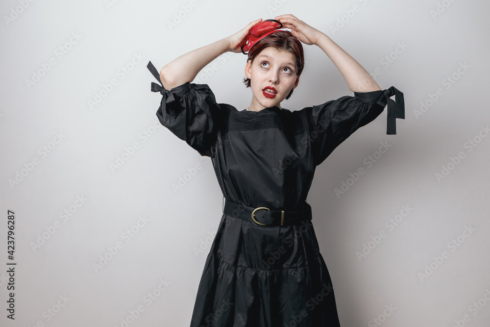 A fashionable beautiful girl in a black dress, straightens her red little hat with her hand. white background