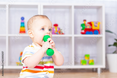 Funny cute caucasian baby boy playing with small dumbbell. Sitting on white carpet in striped bodysuit. Different children's toys in the background. Green dumbbell in mouth. 