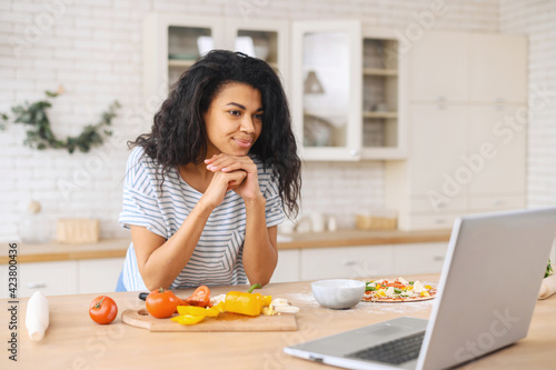 Interested mixed-race girl with hands on chin attentively watching cooking classes, learning how to make dinner lunch watching video blog course from laptop in kitchen, veggies ingredients are ready