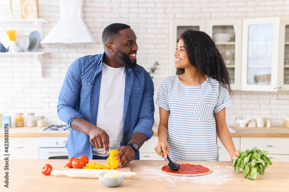Overjoyed black couple standing next to kitchen counter, spending weekend together at home, looking at each other, cooking, girl responsible for spreading sauce on pizza, guy chopping veggies peppers Overjoyed black couple standing next to kitchen counter, spending weekend together at home, looking at each other, cooking, girl responsible for spreading sauce on pizza, guy chopping veggies peppers