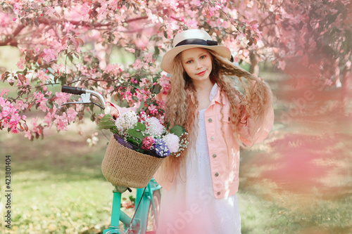 Portrait of beautiful longhaired blonde girl, She wearing pink jacket and straw and walking with her bicycle in apple blooming garden