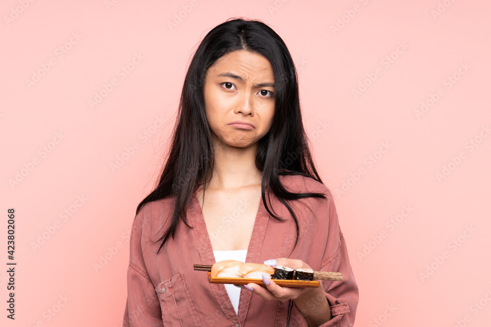Teenager Asian girl eating sushi isolated on pink background sad