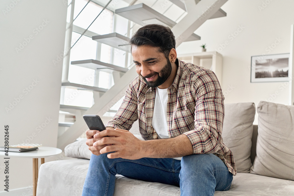Happy young indian man using smart phone sitting on couch at home. Smiling bearded ethnic guy holding mobile phone watching social media video on smartphone, ordering food delivery online or messaging