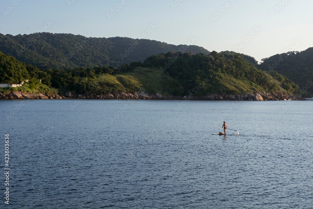 Natural landscape in the port channel of the city of Santos. A standup man padding along with his dog on the board. In the background, the mountains of the city of Guarujá..