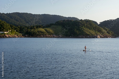 Natural landscape in the port channel of the city of Santos. A standup man padding along with his dog on the board. In the background, the mountains of the city of Guarujá..